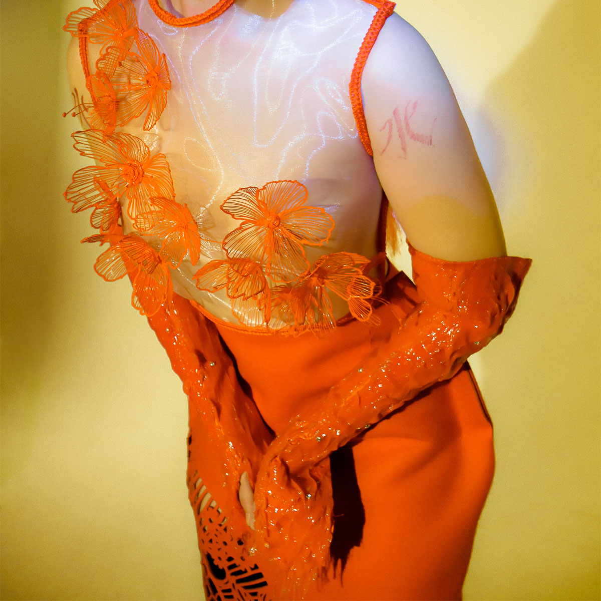 A model is wearing a bright oragne garment with see-through panels and orange vinyl gloves. The model is slightly leading forward with theor hands in front of their lower torso, in front of a yellow backdrop.