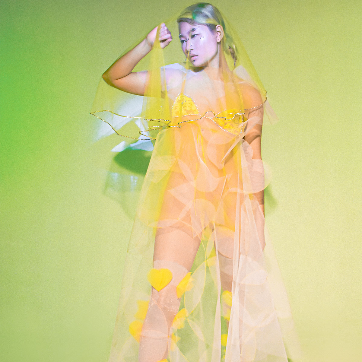 A model is wearing a see-through yellow garment made of krinolin or organza. The model is standing with their hand raised to their face, in front of a bright green photo backdrop.