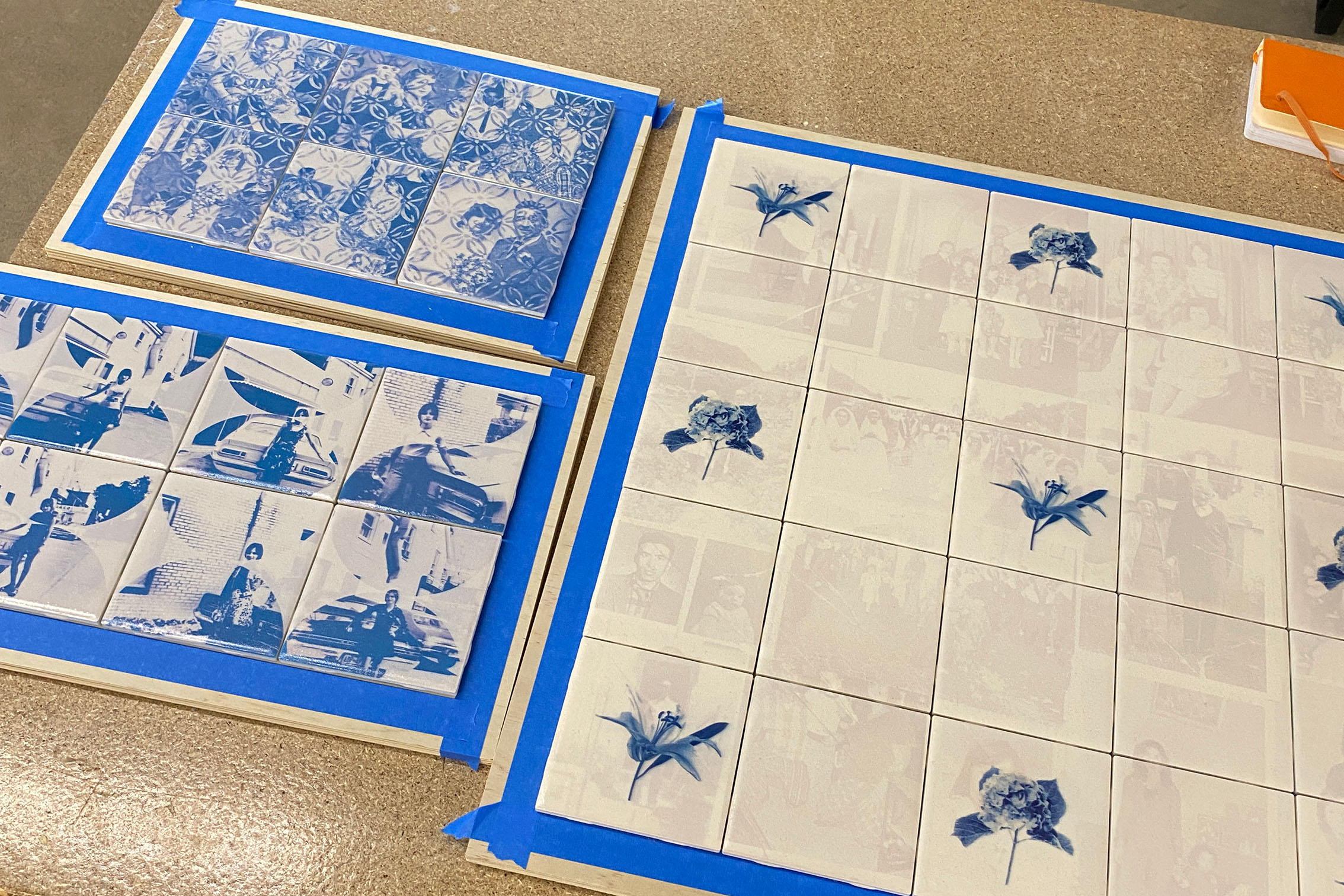 An array of tiles placed on top of boards surrounded by blue painters tape. some tiles have blue images printed on their surface.