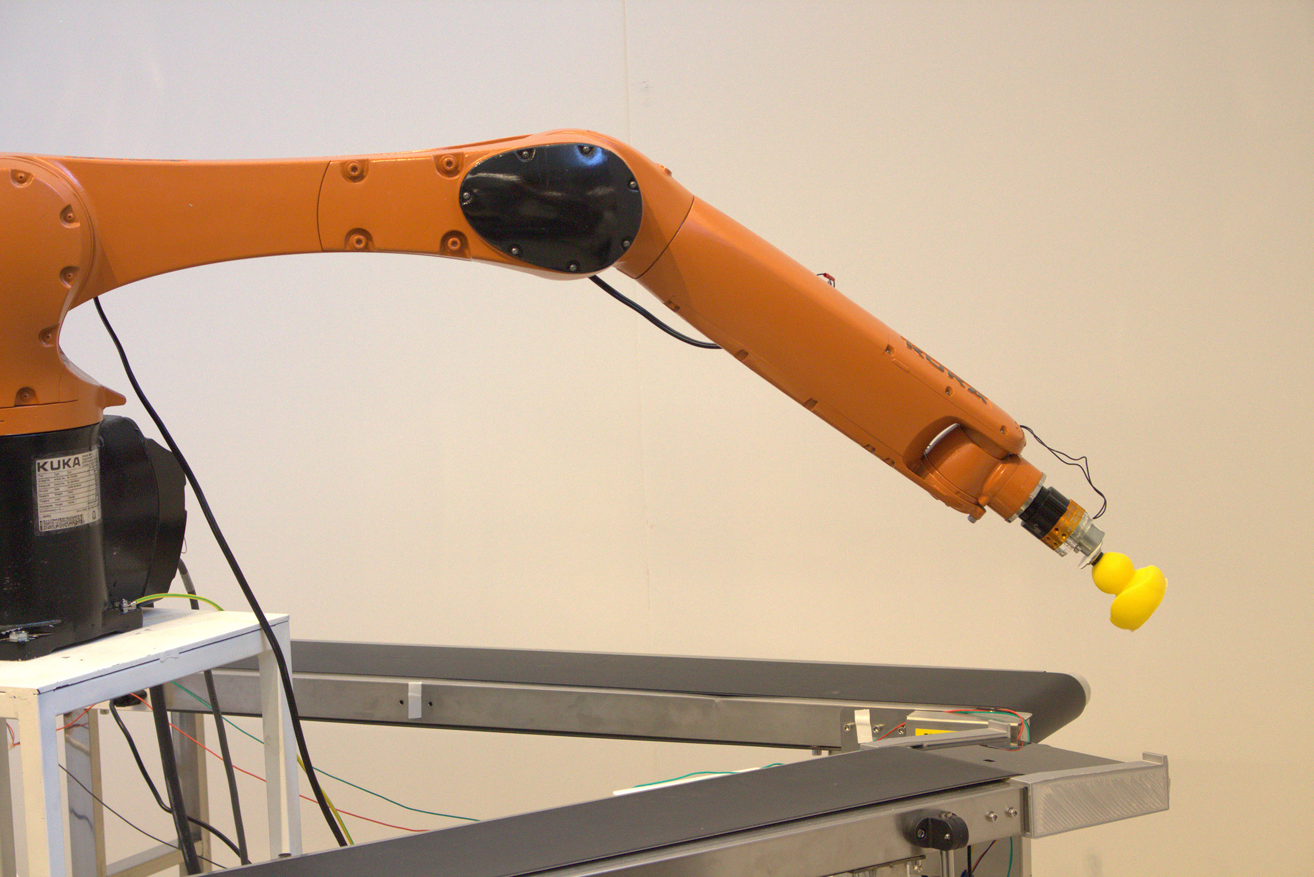 An orange robotic arm is extended and holding a yellow rubber duck over conveyor belts.