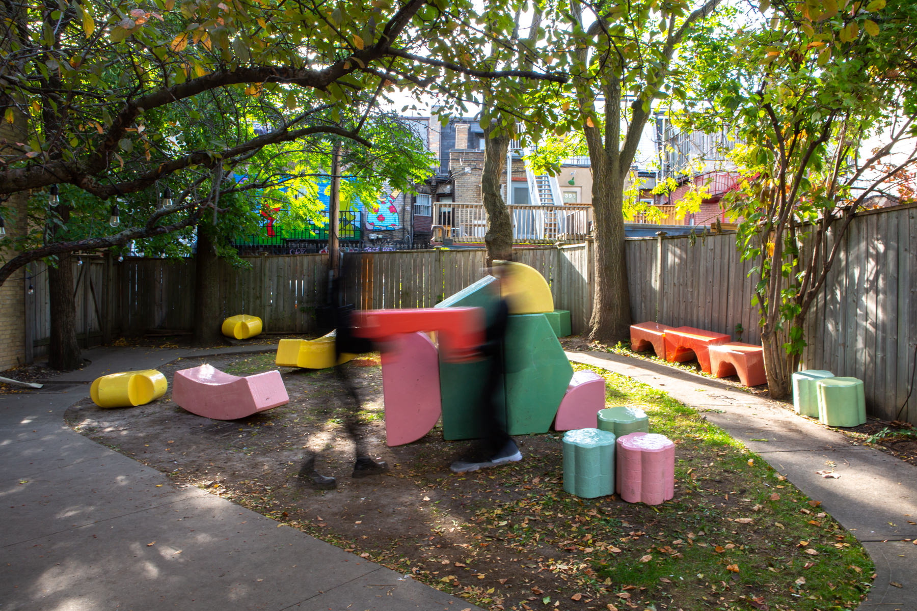 Wide-angle view of about 20 large-scale, colourful blocks from Cecil Community Planting Imagination project. 2 people are photographed moving quickly in front of the blocks.