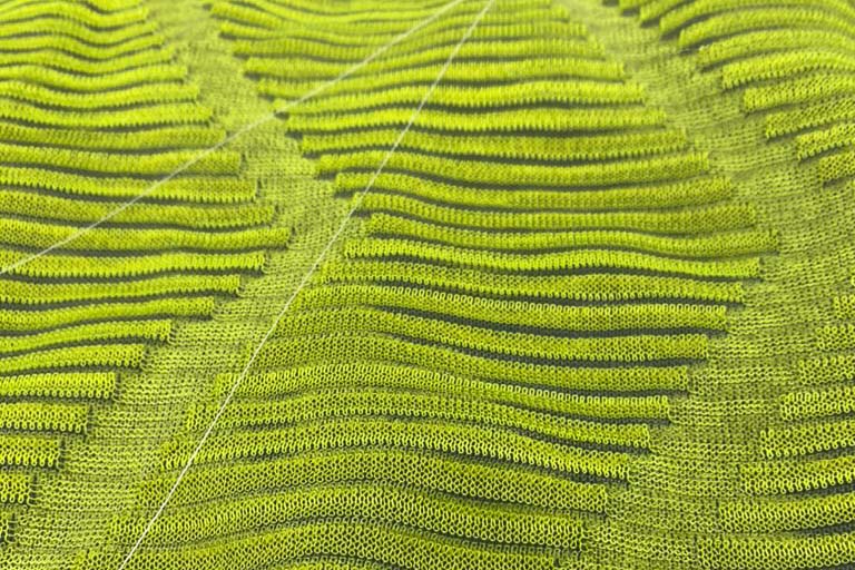 Close-up image of a machine-knit piece. Neon green threads are interwoven with whit and black threads in a herringbone pattern. 