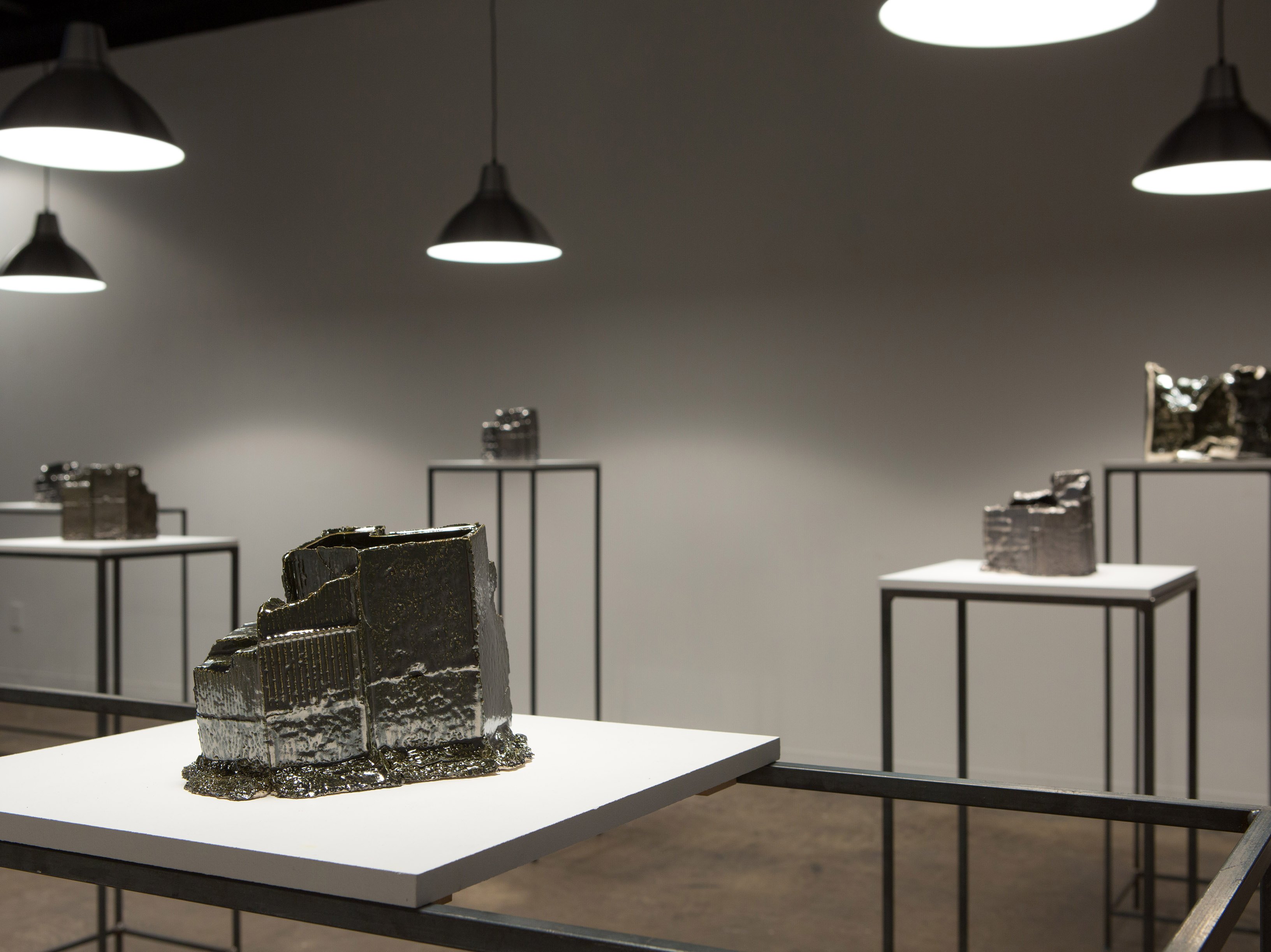 A room full of metallic-coated ceramic forms on plinths. Above each plinth is a low-hanging light fixture. 