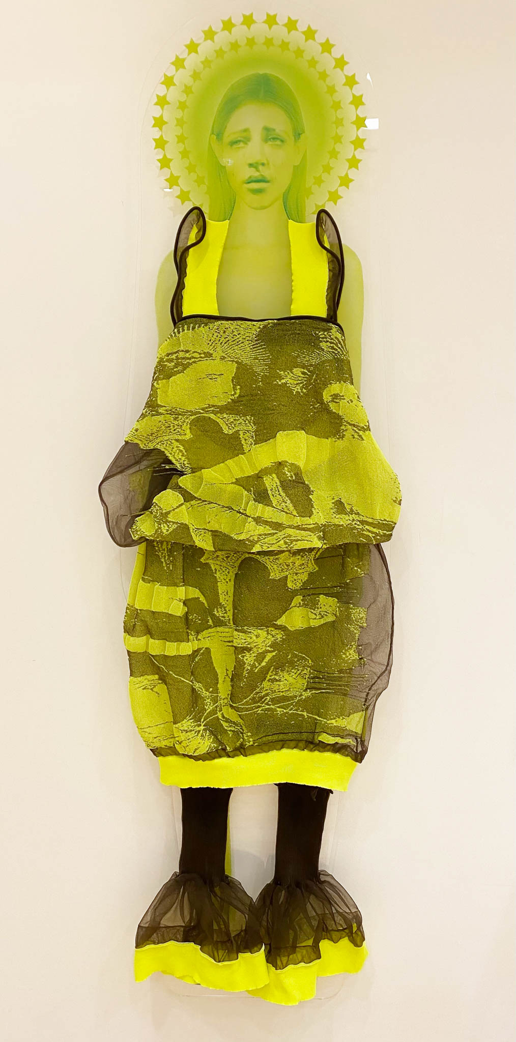 One neon green printed women wearing a ruffled dark grey and neon yellow garment from the Veiling Veronicas series. The piece is mounted to a white wall as if it's floating.