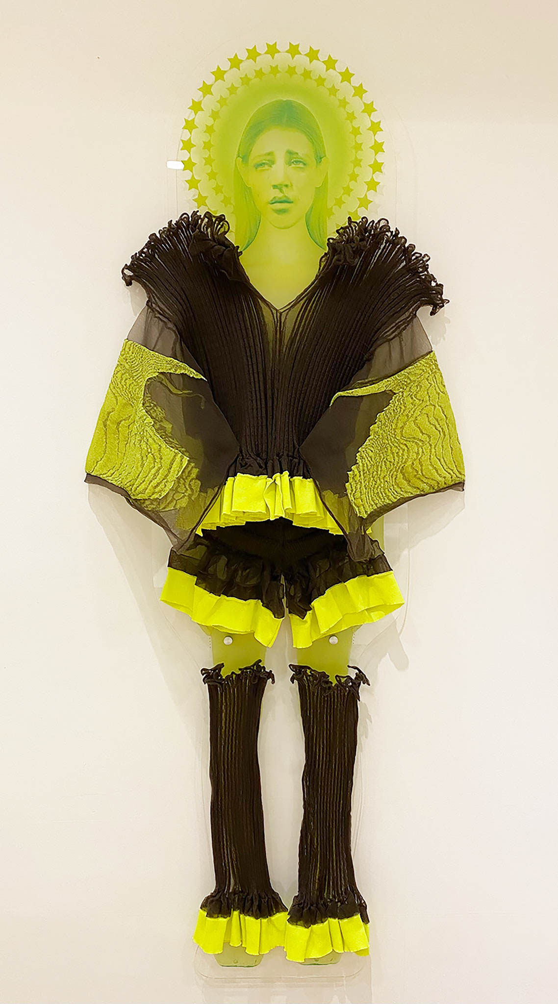 One neon green printed women wearing a ruffled dark grey and neon yellow garment from the Veiling Veronicas series. The piece is mounted to a white wall as if it's floating.