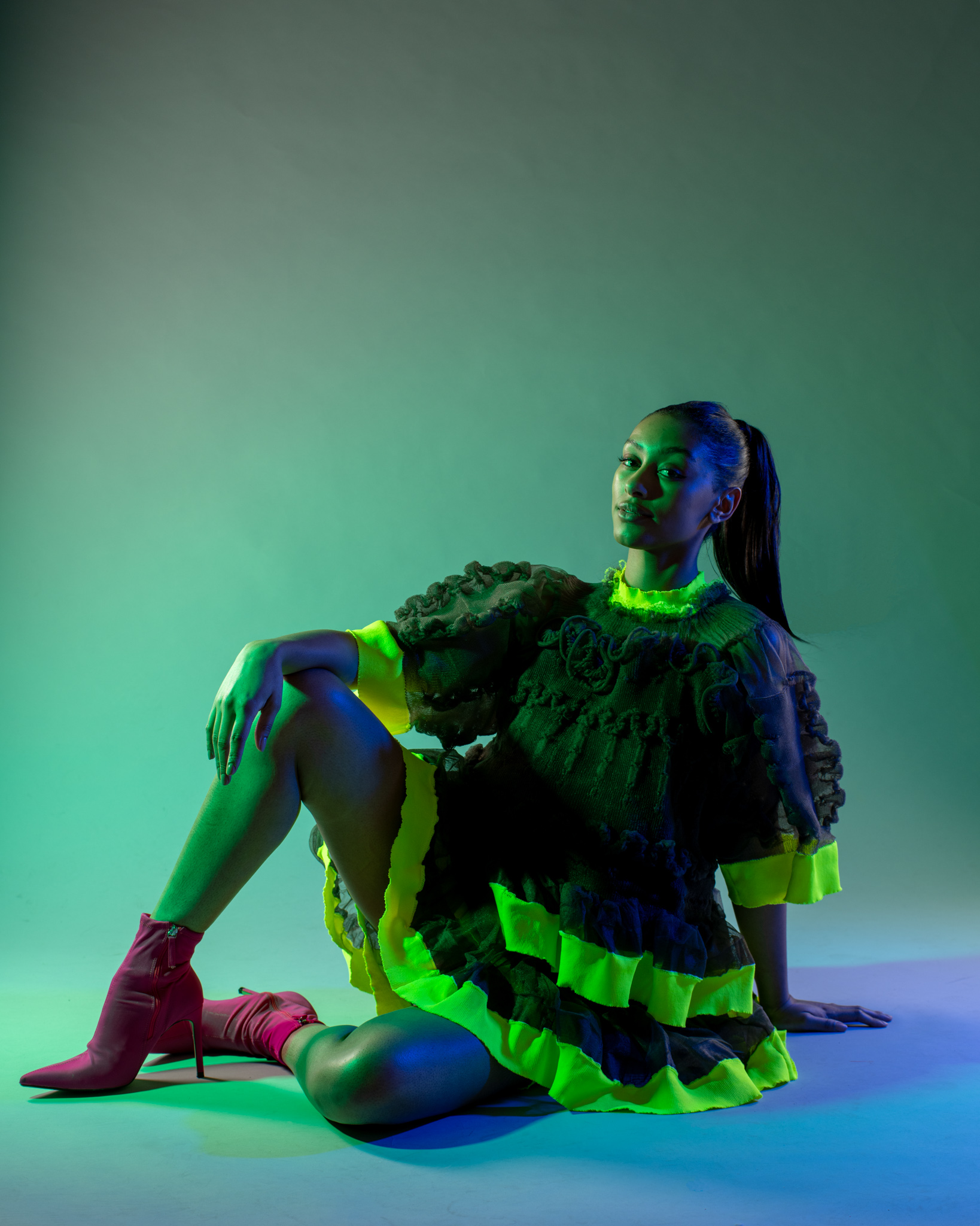Fashion model posing on the ground with knee raised up. Green lighting. Showcasing one of the garments from Veiling Veronicas series.