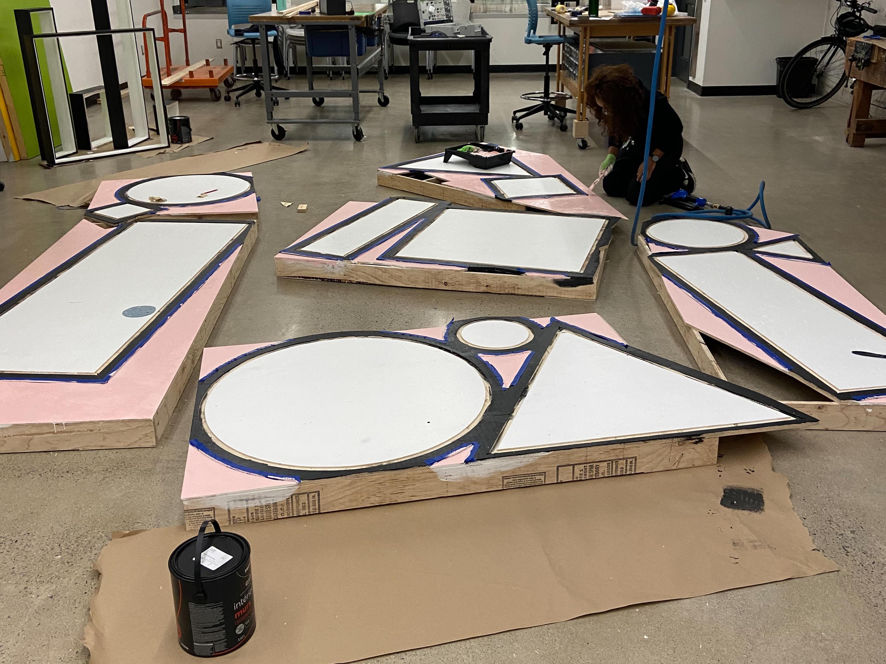 Large wooden shapes are spread out on the floor of a workshop. They are being painted white, black and light pink.