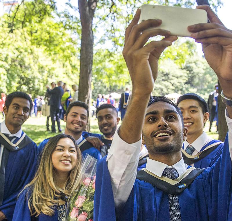 A group of diverse graduating students taking a selfie while wearing their graduation robes outside in the Quad. 