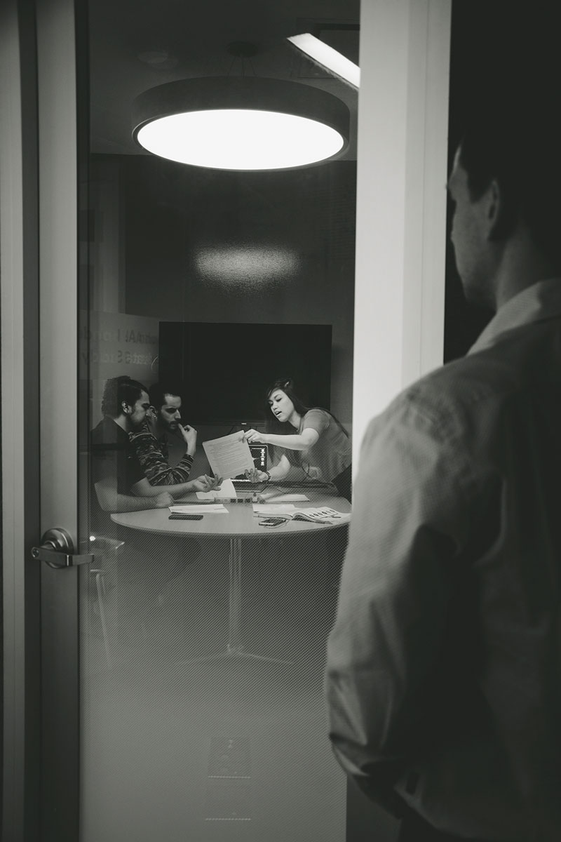 Man looking into a room from a doorway at 3 people sitting at a round table talking and looking at papers together. 