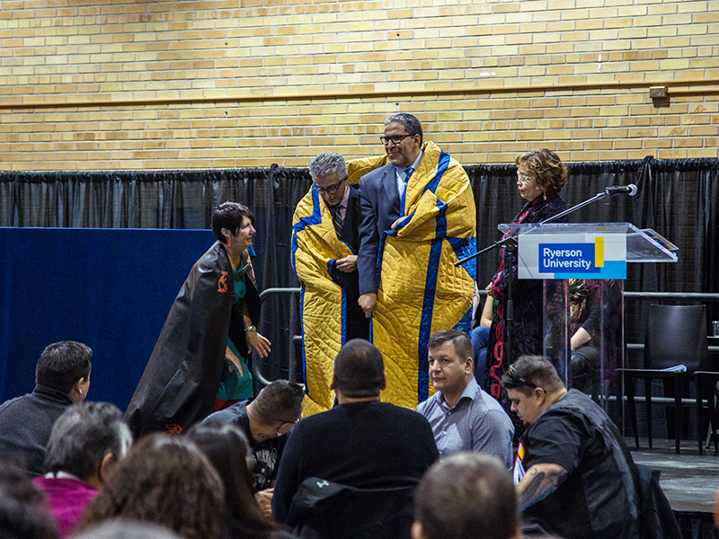 Michael Benarroch and Mohamed Lachemi being wrapped in the yellow and blue star blanket at the Truth and Reconciliation at Ryerson community celebration