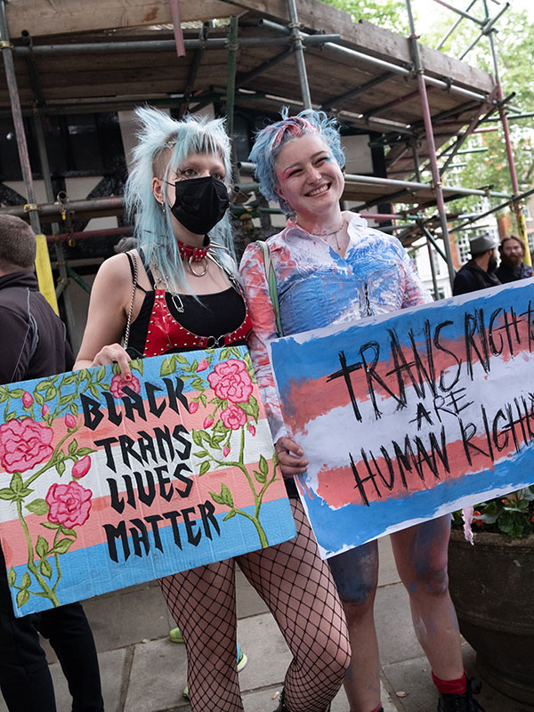Two people posing for the camera holding signs stating. Black Trans Lives Matter, Trans Rights are Human Rights.