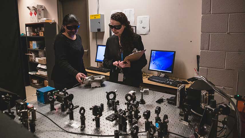 A member of the Environmental Health and Safety team and a Ryerson staff member review the components in a laser lab while wearing dark protective glasses.