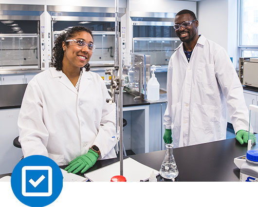 Two students working in a chemistry lab on campus while wearing personal protective equiment.