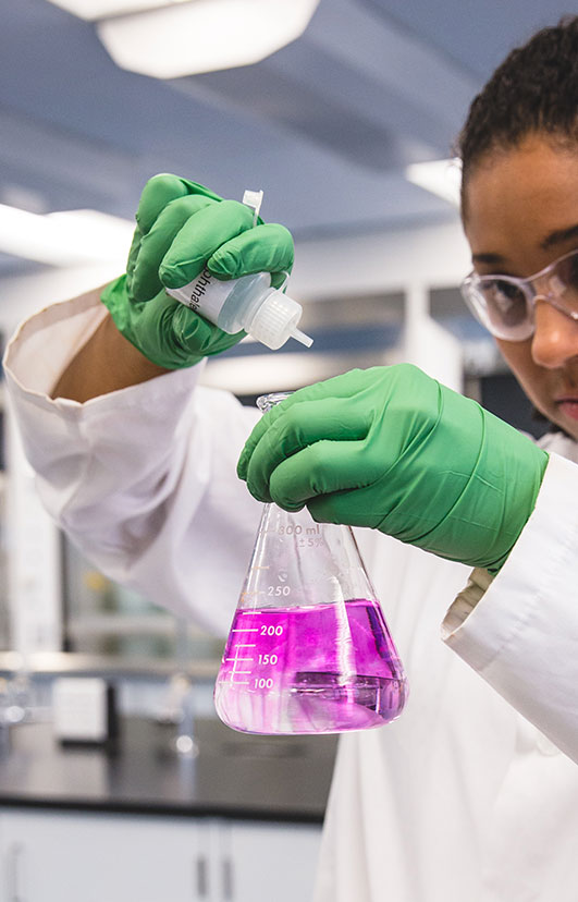 A student wearing green rubber gloves, dropping a solution into a flask containing a pink liquid. 