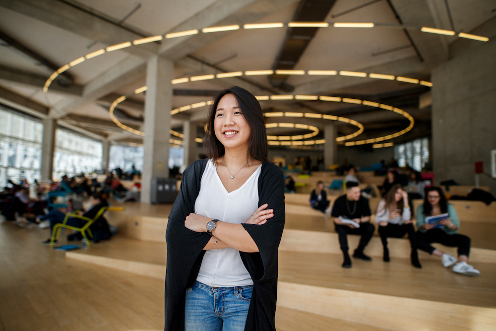 A female student is standing at the Student Learning Centre, smiling and looking away from the camera. Students can be seen in the background.