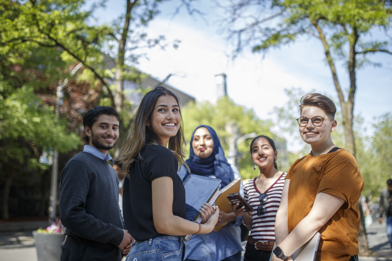 A group of students are hanging out with each other on campus. Four of them are smiling and looking off into the distance while the fifth student is looking at the camera.