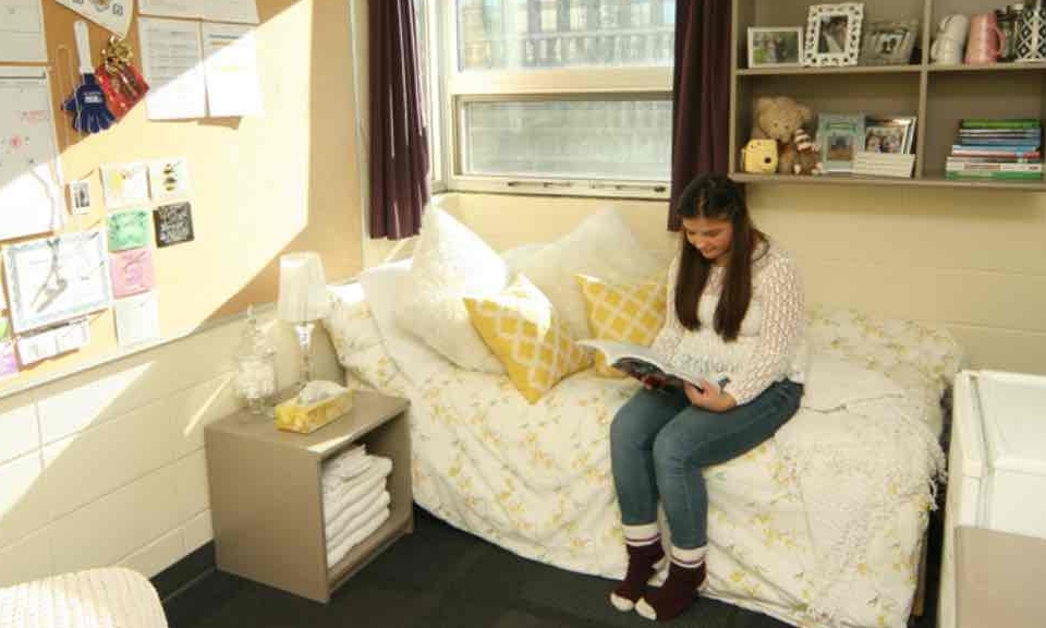 A student is sitting on her bed in a dorm room reading a book. 