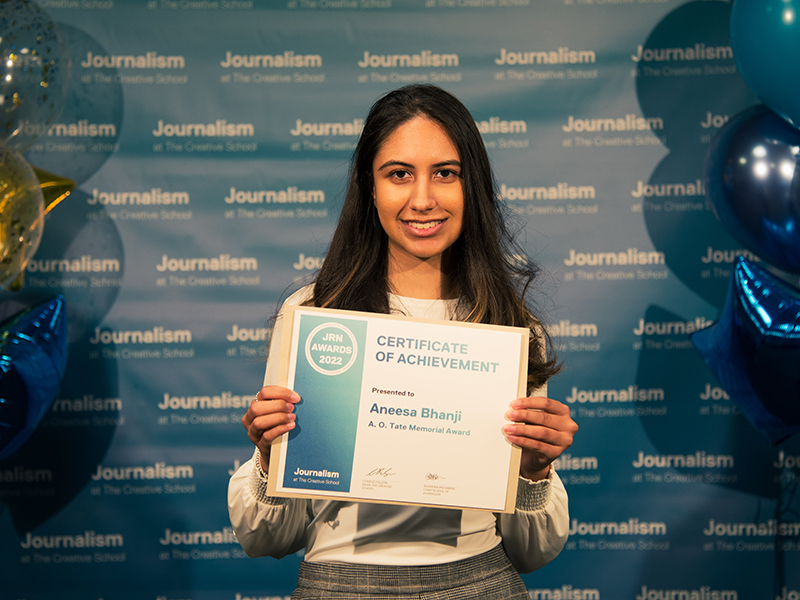 Aneesa Bhanji holds her certificate while standing in front a blue backdrop that repeats, "Journalism at The Creative School."
