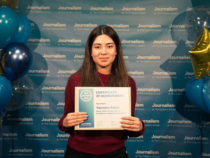 Yasmeen Aslam holds a certificate while standing in front of a blue backdrop that repeats, "Journalism at The Creative School."