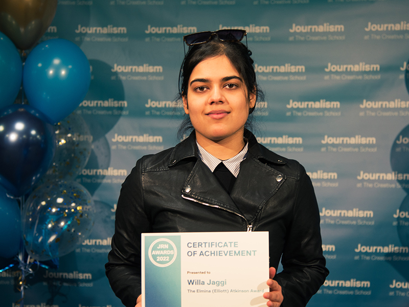 Willa Jaggi holds a certificate while standing in front of a blue backdrop that repeats, "Journalism at The Creative School."