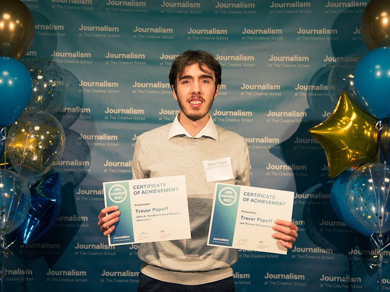 Trevor Popoff holds two certificates while standing in front of a blue backdrop that repeats, "Journalism at The Creative Schoool."