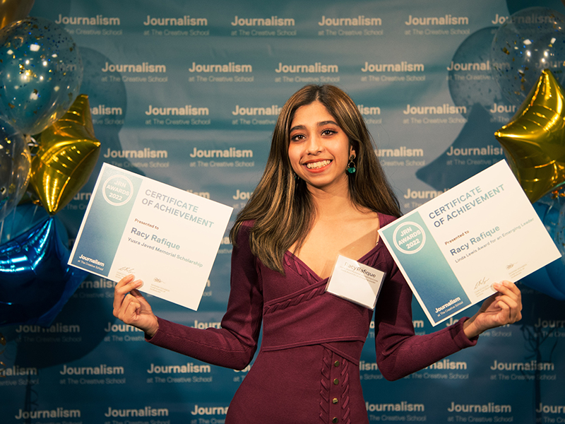 Racy Rafique holds two certificates while standing in front of a blue backdrop that repeats, "Journalism at The Creative School."