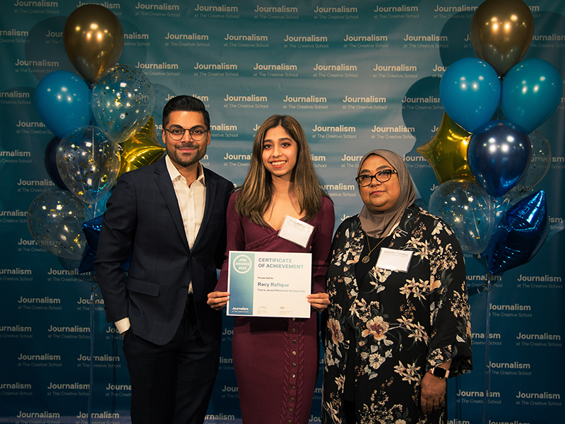 Racy Rafique holds a certificate while standing with Colin D'Meilo and Sabrina Suraiya in front of a blue backdrop that repeats, "Journalism at The Creative School."