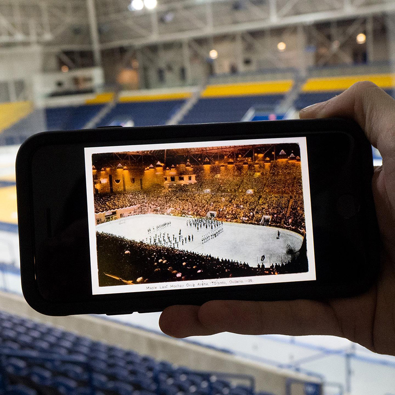 In the empty Mattamy Athletic Centre, a phone is held up with an image of a past hockey game with a full crowd