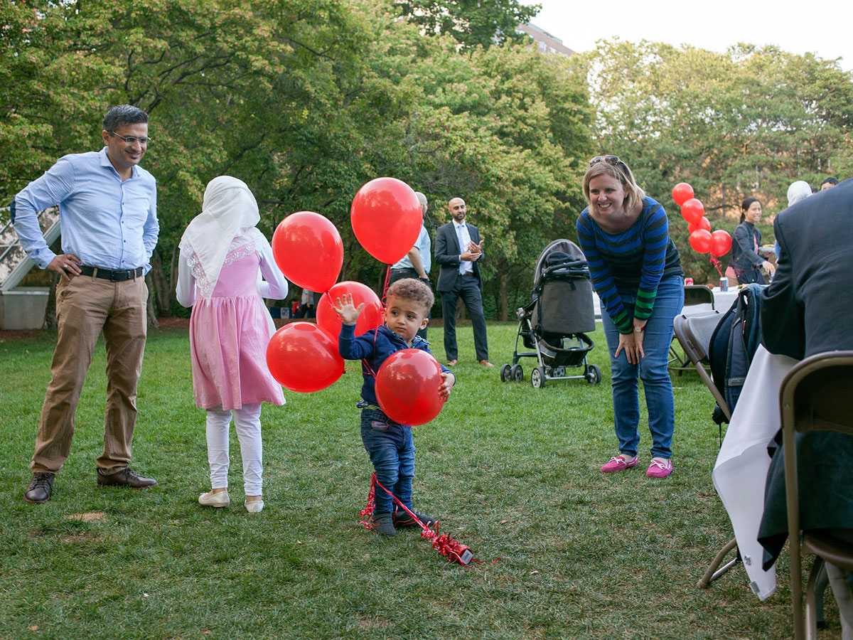 A little boy holding red balloons with adults talking behind him