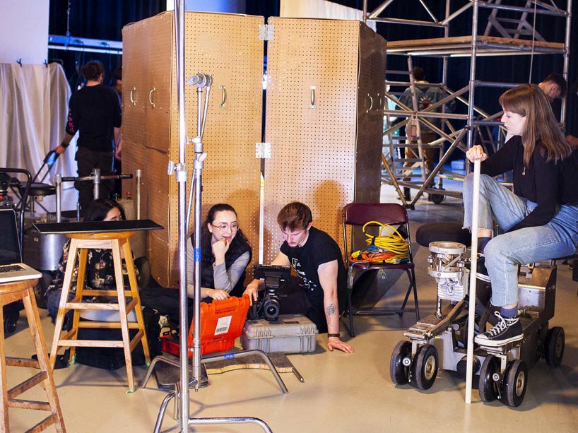 Three students sitting on the floor, a fourth sitting on machinery, looking at film scenes on a camera