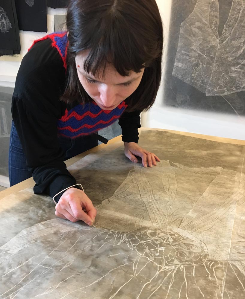 Artist Sarah Casey is bending over her drawing of a 1927 wedding dress, laid out on a work table, etching lines into the waxed paper with a dressmaker’s pin