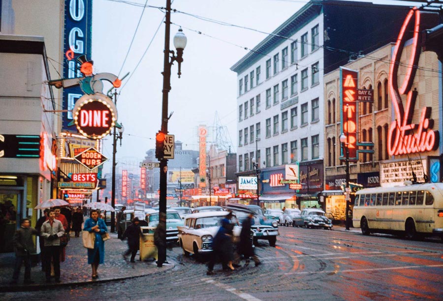 Colour photograph of a corner on Vancouver’s Granville Street in 1959, with pedestrians crossing the road in front of stopped traffic. Parked cars and buses also line the road, and neon restaurant, theatre and store signs that read “Studio,” “Dodek Furs,” “Best’s Ladies Apparel,” “Bowling,” and “DINE” shine on both sides of the street