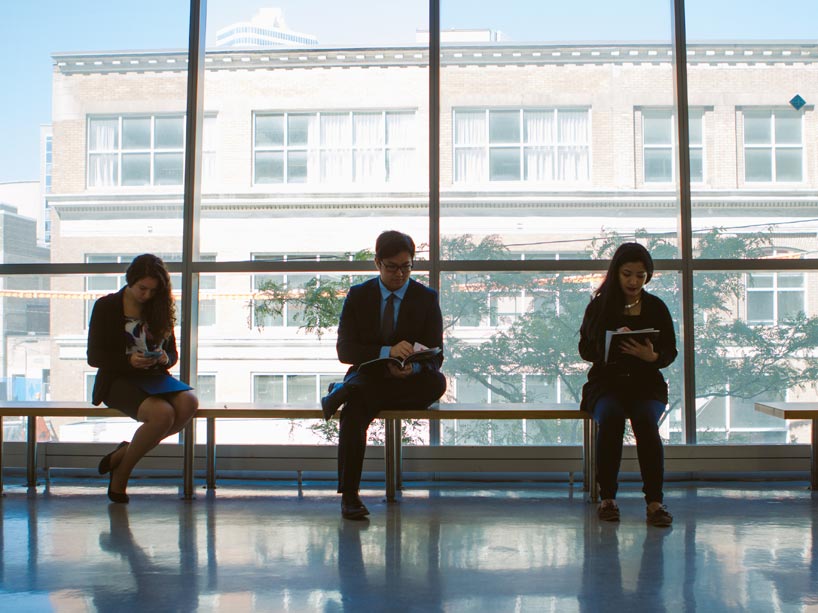 Three students dressed in business attire, sitting on a bench and looking at their phones and portfolios
