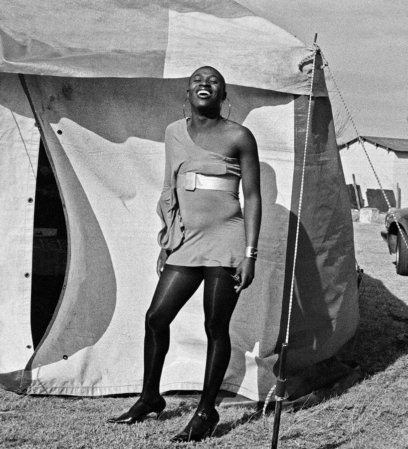 A black and white image of an African transwoman laughing and standing in front of a tent