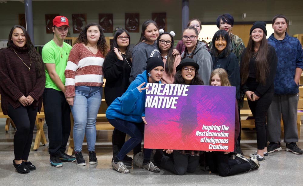 Creative Native participants pose with program founder, Buffy Sainte-Marie, in front of a sign with a logo they designed