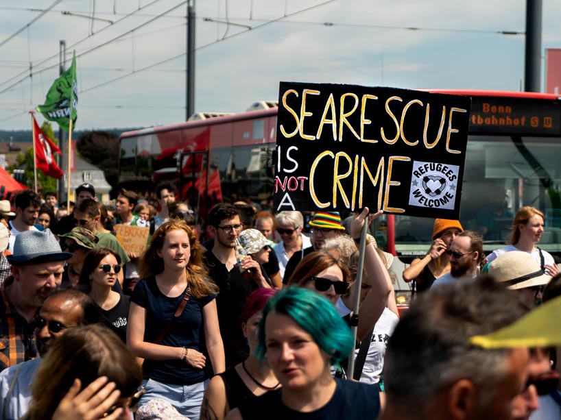 Protesters marching, one holds sign reading “sea rescue is not a crime, refugees welcome”