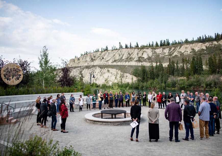 People standing in a circle around a fire pit with a wooden Indigenous sculpture to the left”