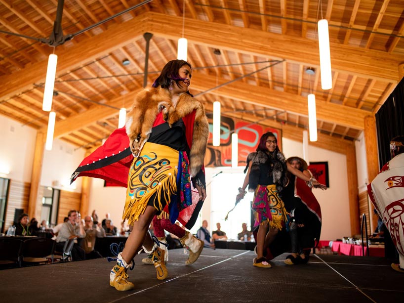 Four Indigenous females wearing traditional regalia dance on stage