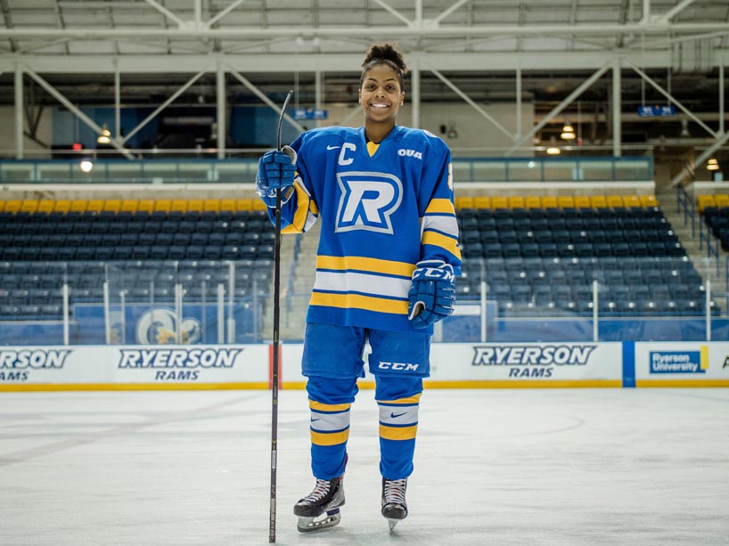Female hockey player wearing full uniform, standing on ice in her skates, holding her hockey stick