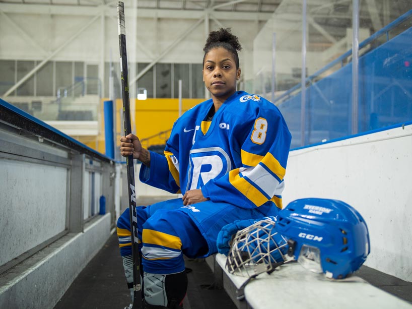 Female hockey player wearing full uniform sitting on the team bench, holding her hockey stick