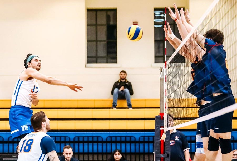 A male volleyball player jumps to spike the ball over the net while two opposing players block