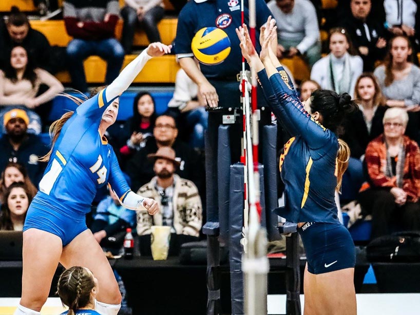 A female volleyball player jumps to spike the ball over the net while an opposing player blocks