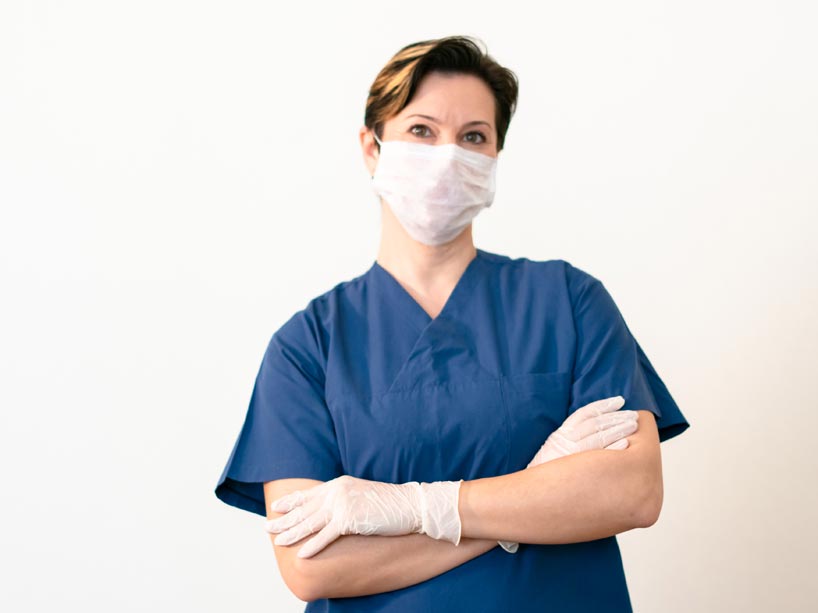 A person in scrubs stands with crossed arms wearing gloves and a medical mask