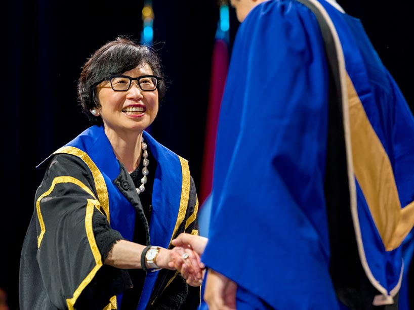Janice Fukakusa on stage at convocation dressed in a black and blue gown shaking hands with a graduate