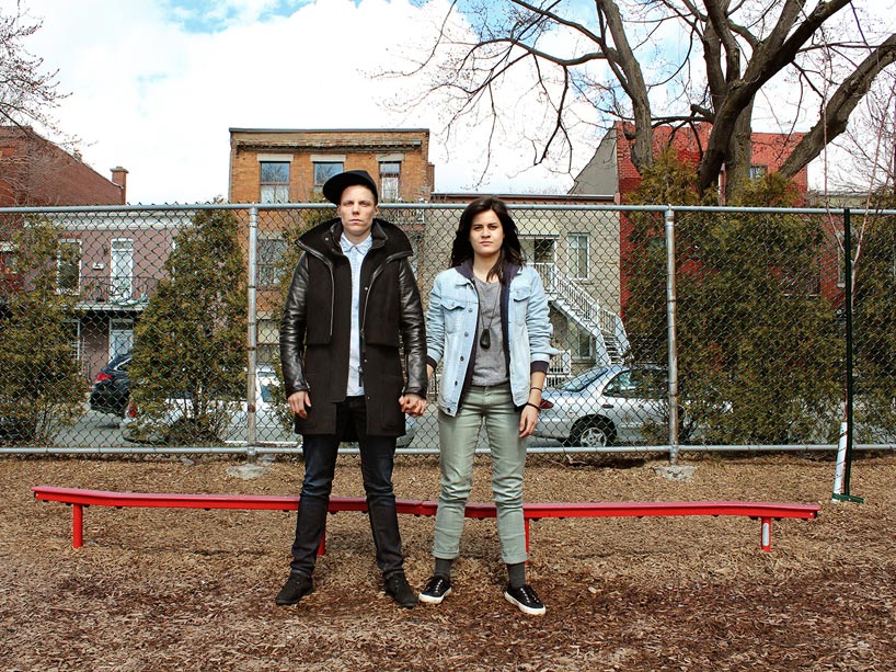 Two young people stand side by side, hand in hand in front of a low red bench with a chain link fence in the background