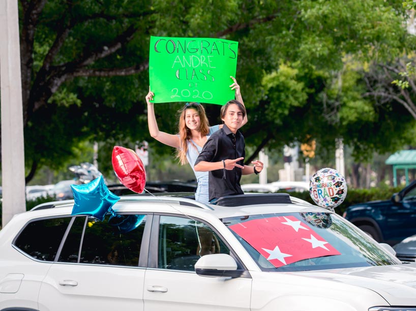 Two people standing up through the sunroof of a parked car holding congrats signs
