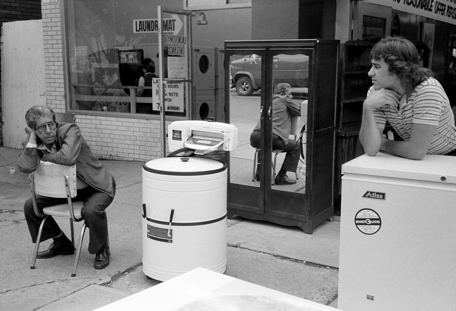 Two men sit outside on the street with a wringer washer between them