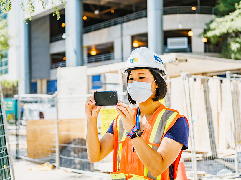 A woman on a construction site wearing a hard hat and mask taking a photo with her cell phone