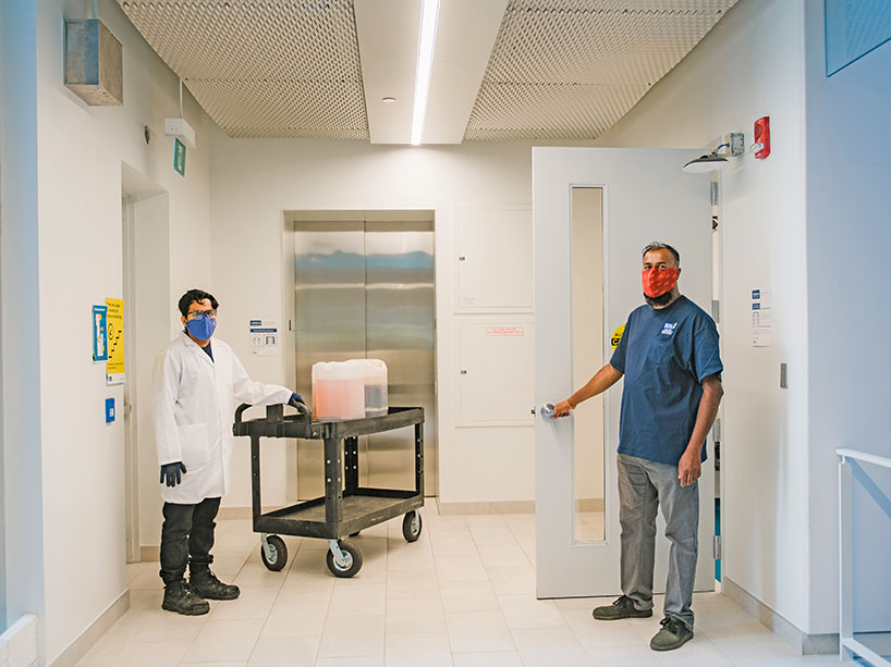 Two men wearing masks, standing away from each other, in a hallway