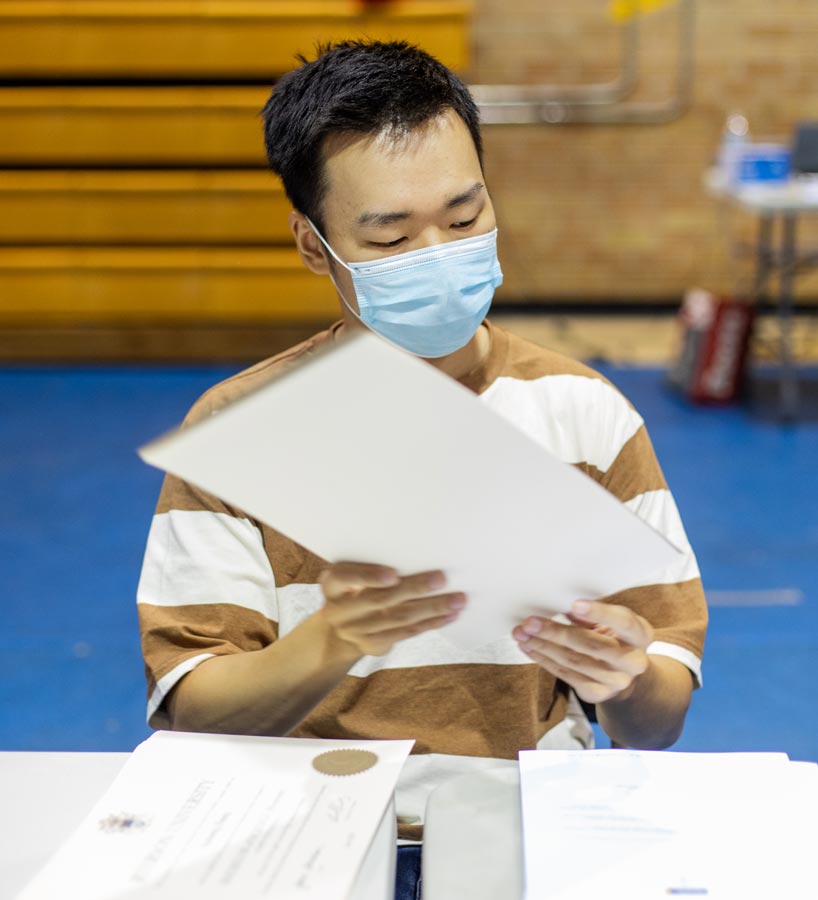 Seated male student assembles packages of paper while wearing a face mask